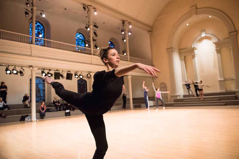 A female ballet dancer executes a high arabesque while the ensemble forms poses in the background on Danspace's steps
